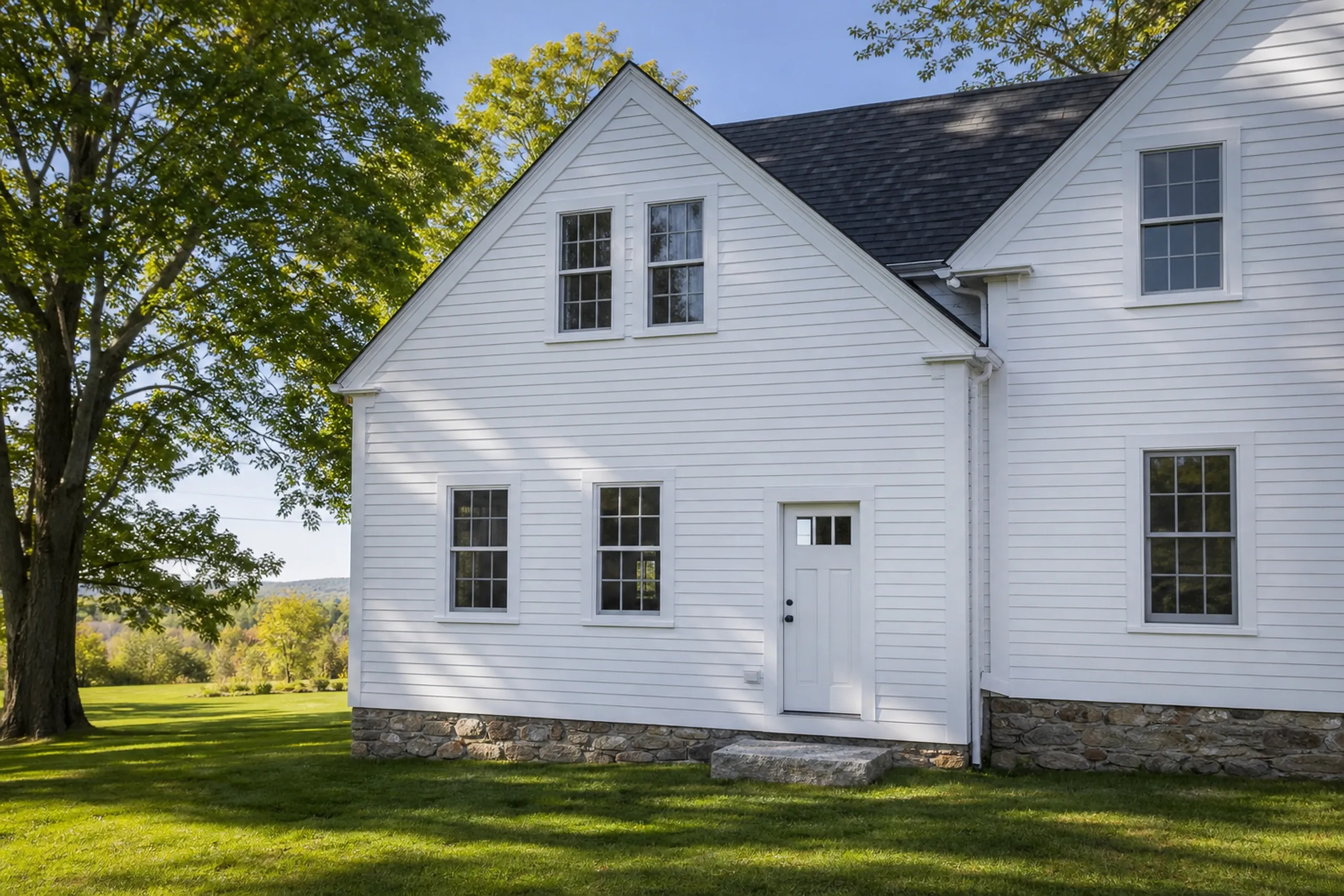Finished home addition near Bangor, ME: white farmhouse with dark roof, stone foundation, multiple windows, white trim, large tree in yard.