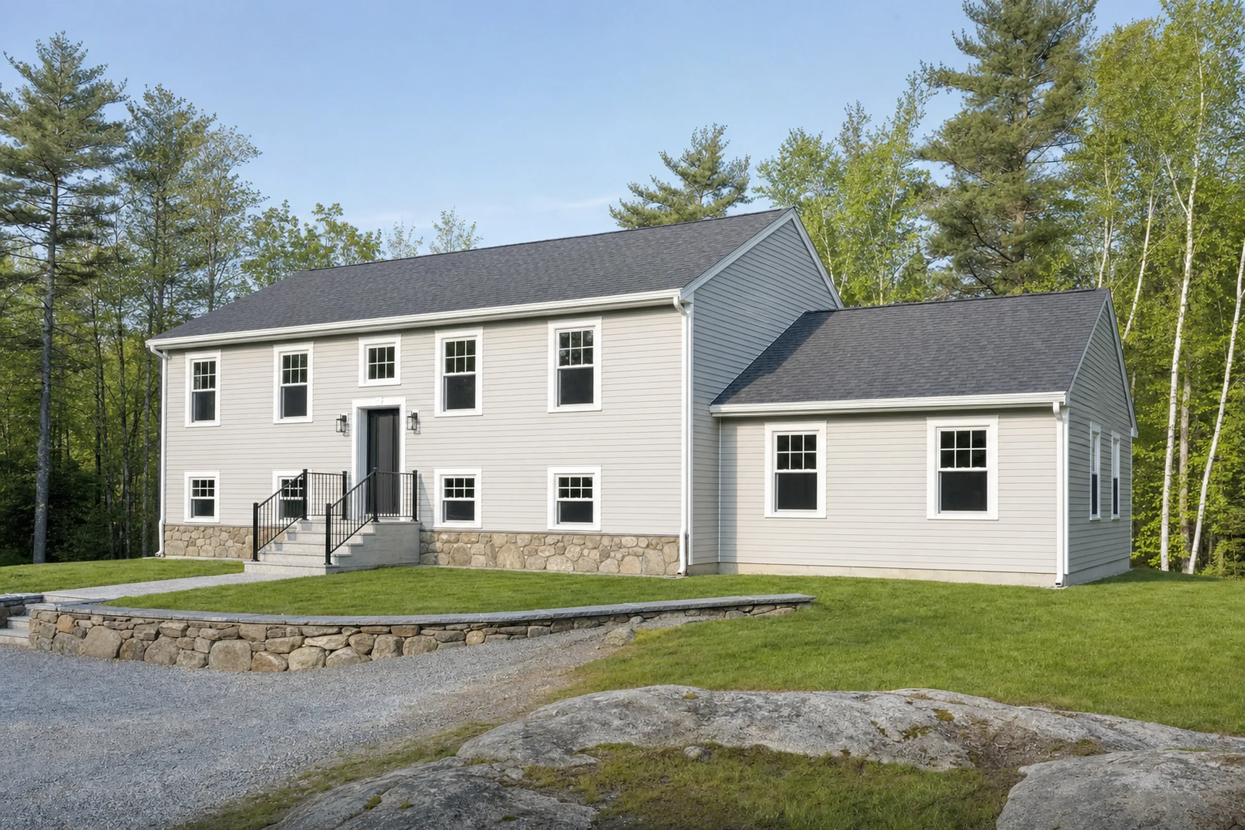 General contractor in Holden, ME: Two-story white vinyl-sided house with dark gray roof, black trim, stone foundation, metal railing, surrounded by trees and grass with gravel driveway.