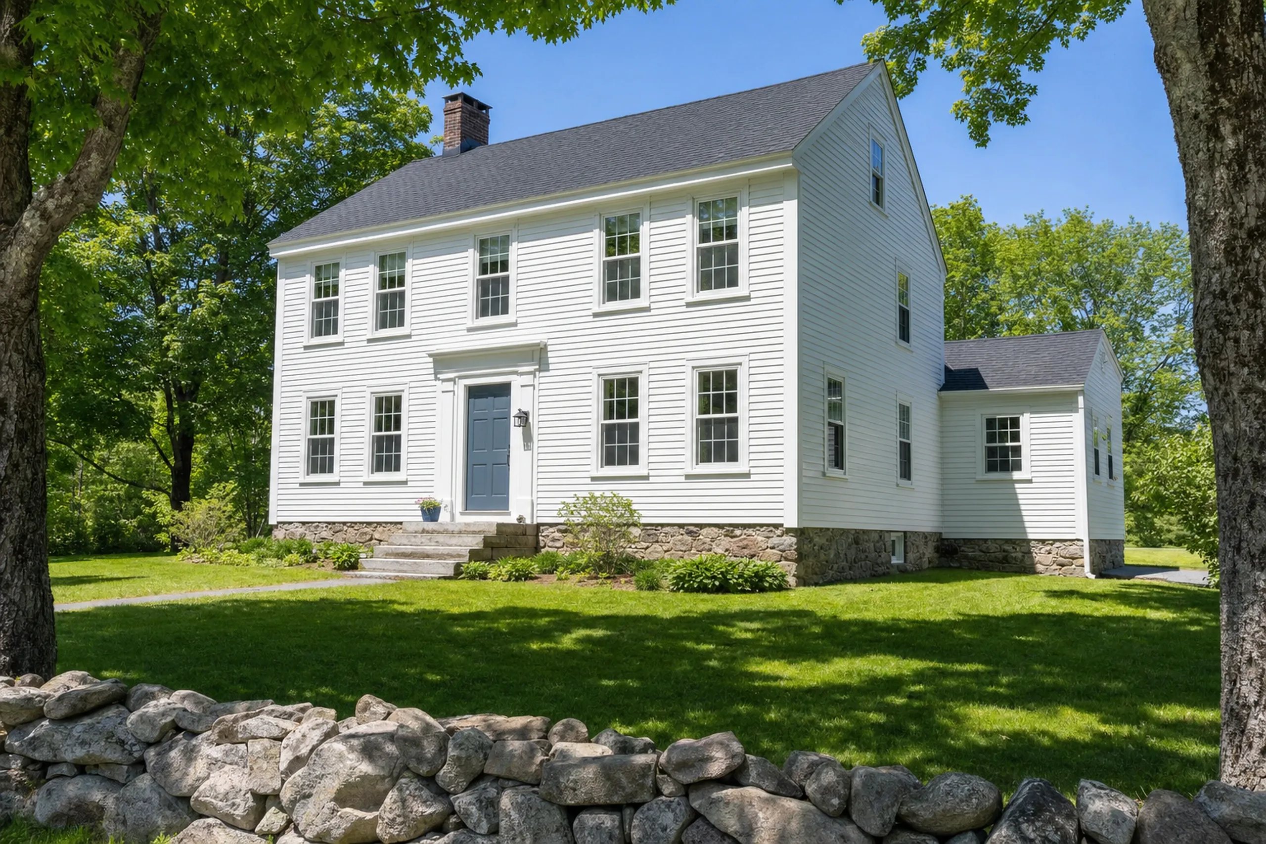 General contractor in Old Town, ME: White two-story colonial house with white vinyl siding, black shutters, and gray roof surrounded by green lawn and large trees.